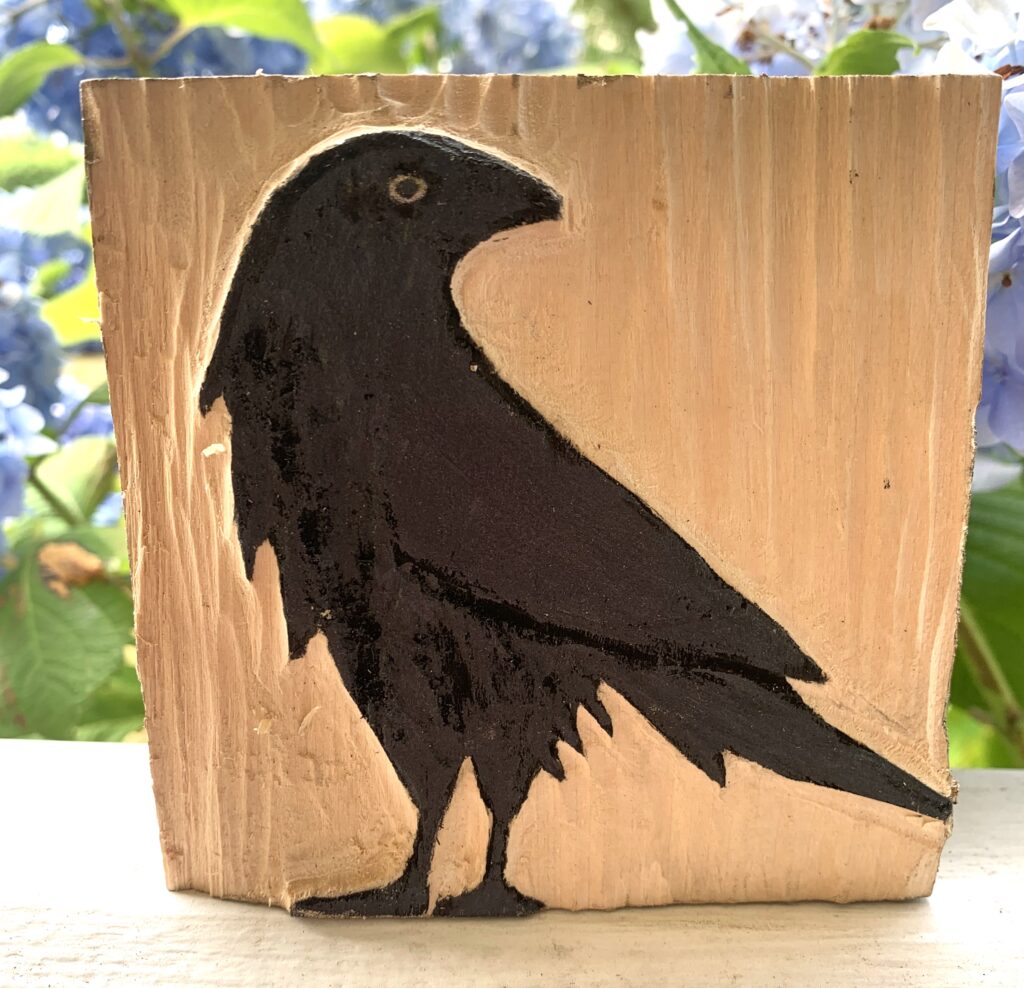 Black crow profile carved in wood block photographed in front of hydrangea